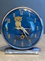 Front view of the round owl alarm clock with blue dial, gold numerals, and silver casing on stand.