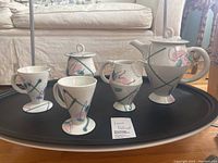 Full set of tea service items arranged on black tray including teapot, sugar bowl, creamer, two teacups, and artist card showing Laurie Rolland name and Toronto address.