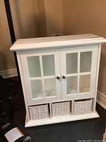 White manufactured wood bathroom cabinet with glass panel doors and 3 wicker baskets at the bottom, shown standing on floor against wall.