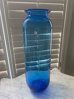 View of tall blue Blenko glass vase showing shape and color, standing on marble surface in front of window blinds.