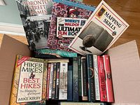 Top view of the box showing a collection of books including Toronto Street Names, Where's Waldorf Ultimate Fun Book, Mapping Skills, and Hiker Mike's Best Hikes, with DVDs lined up beside them.