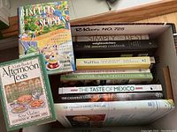 Overview photo of 11 cookbooks in a box, titles visible and showing a variety of cooking topics such as biscuits, scones, afternoon teas, waffles, Mexican food, and Canadian living themed cookbooks.