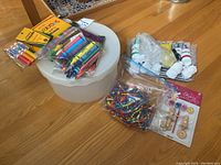 Overview photo showing round plastic box with clear lid, packets of assorted crayons and markers, plastic beads, small paints, and ceramic figures on wooden floor.