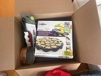 A cardboard box containing the chip maker and mandoline box, the bottle brush handle, and the copper saucepan