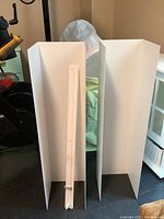 Two white metal IKEA shelves with U-shaped bent vertical panels and one mounting bar leaning against a shelf.