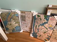 Three framed Plak-it maps lined on floor including Brooklyn, Midtown Manhattan, and NY Subway maps, wrapped with corner protectors and plastic sealing.