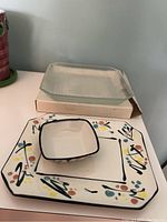 Photo shows square ceramic decorative plate with colorful abstract floral pattern and smaller matching bowl placed on it, along with stacked glass plate in box behind.