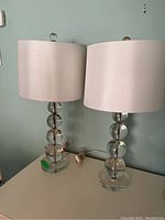 Two glass base table lamps with white fabric drum shades shown side by side on a white surface against a light blue wall.