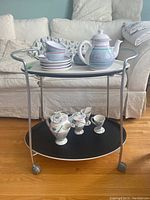 Tea cart side view showing white tray on top with ceramic tea set and black tray below.