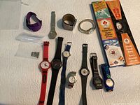 Overview of 11 vintage collectible watches laid out on a white quilted surface including wristwatches of various styles and colors, two McDonald's official Olympic watches in original boxes, and a few extra metal links.