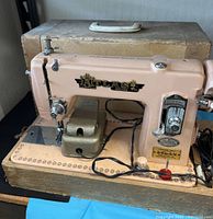 Vintage Atlas Precision Deluxe sewing machine with attached foot pedal, pink painted metal body, visible branding, on top of worn beige carrying case