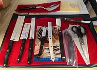 Full layout on red mat showing knives, manuals, utensils, honing steel in sheath, polishing cloth and case