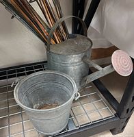 Photo showing galvanized steel bucket and watering can on metal shelf, displaying shape, size, and minor surface oxidation.