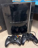 Sony PlayStation 3 console with PlayStation branding and four wired controllers placed in front with cables wrapped around them.