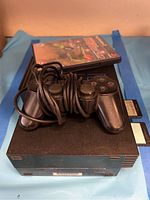 Sony PlayStation 2 console with controller and game shown stacked on blue cloth.