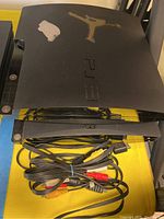 Two black Sony PS3 consoles stacked with AV cables, showing top console with stickers and wear.