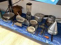 Photo showing various vintage cookware and kitchen items laid out on a blue tarp including a copper pot, blender, metal pans, mechanical kitchen tools, and a funnel.