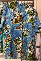 Front of vintage Hawaiian shirt showing detailed tropical island print with surfers, palm trees, and ocean waves. Wooden style buttons and a chest pocket visible.