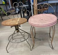 Photo showing two vintage vanity stools side by side on floor, one with wooden swivel seat and one with pink cushioned seat, both with decorative metal scrollwork backs.