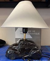 Black ceramic Volkswagen Beetle car shaped table lamp with white fabric shade and original box in background.