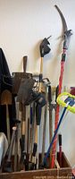 Photo shows a collection of yard tools standing up against a wall, including shovels, rake, manual pruning saw, and garden shears, some with wooden handles and metal blades.