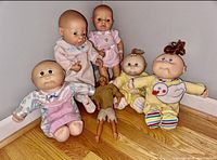 Photo showing a group of dolls: 1 Ken Doll, 3 Cabbage Patch Dolls, 2 Berjusa rubber babies arranged on floor with wooden background.