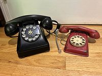 Black antique rotary telephone and red metal toy rotary telephone placed side by side on wooden floor, showing both items fully