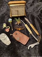 Full view of all items on cloth backdrop: clock, leather key holder, keys, heart lock, stick pin, brooches, cutlery, and keychain.