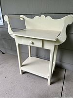 Full view of off-white painted farmhouse washstand with single drawer and curved back yoke, lower shelf visible.