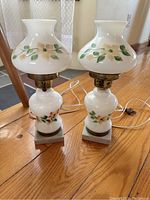Pair of white milk glass hurricane lamps with floral painted designs captured standing on wooden floor. Lamps feature brass fixtures and square marble bases.