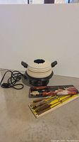 Photo of white ceramic fondue pot with black and yellowish stripes, two handles, electric base and cord, and a box of fondue forks placed on the floor.
