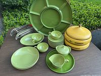 Full view of vintage green melamine dishware set including plates, cups, bowls, oblong bowl, sugar bowl with lid, cream pitcher, along with yellow canisters, Lazy Susan, and french fry cutter on table.
