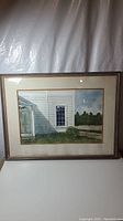 Framed watercolor painting of white house exterior with door, window, and greenery against a partly cloudy sky, shown fully in frame.
