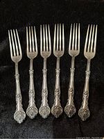 Front view of six matching silverplate dinner forks arranged side by side on a black fabric background, showing detailed pattern and condition.