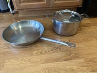 Side view of stainless steel fry pan with long handle and 8 qt stock pot with glass lid on kitchen floor.