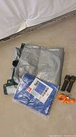 Overview photo showing large silver-gray tarp, blue tarp in packaging, two flashlights, and orange straps on concrete floor.
