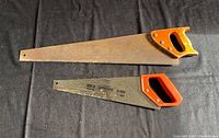Photo showing two manual hand saws with different blade lengths and handle styles lying flat on a dark surface.