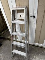 Photo of a metal 5 foot step ladder with white paint, showing wear and paint marks, standing on concrete against a beige wall with two white doors in the background.