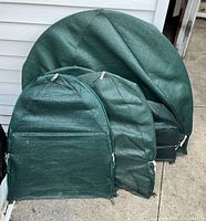 Five green mesh shrub covers of varying sizes displayed against white siding and concrete, showing material and shape details.