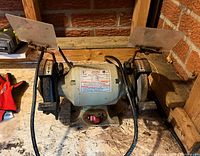 Full view of the Brico bench grinder showing the entire machine with both grinding wheels, tool rests, safety shields, power switch, and power cord.