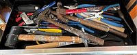 Drawer filled with multiple assorted hand tools including hammers, pliers, utility knives, and a rubber mallet showing signs of use.