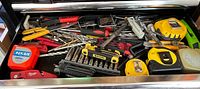 Drawer filled with assorted hand tools including several tape measures, screwdrivers, drill bits, hex key set, and rotary cutter.