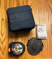 Photo showing laser level with mounting bracket, instruction manual, and black Ryobi carrying case on wood floor