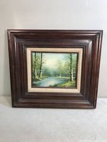Full view of the framed creek landscape painting showing lush trees along a water body, signed C. Tranger and mounted in a dark wood frame with cream edging.