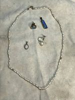 Five jewelry items arranged on a soft fabric: pearl necklace, single opal stud earring in gold, single pearl earring, white opal oval pendant, and blue opal rectangular pendant.