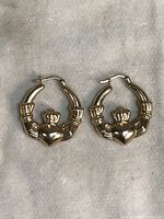 Pair of gold hoop earrings with Claddagh symbol on beige fabric background.