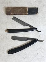 Photo showing two vintage straight razors laid out flat, one with a broken wooden housing and black blade, and the other a black handled razor with metal blade.