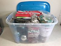 Plastic tub with broken blue lid containing assorted Christmas bulbs, holiday-themed gift buckets, tubs and packaging, including silver bulbs and red and green paper items visible inside.