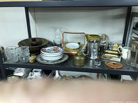 Shelf view with kitchen and collectible items including glassware, ceramic plates, brass perpetual calendar (not working), metal trays, and various containers.