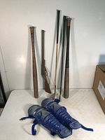 Photo showing two vintage wooden baseball bats, two vintage golf clubs, vintage child's toy rifle, and a pair of blue children's shin guards arranged upright and on table